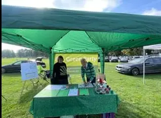 A green gazebo with a Green Party banner on a grass field, and two people at a table of leaflets beneath it.