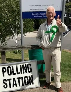 Edward Gildea, wearing a green rosette, giving a thumbs-up beside a polling station sign.