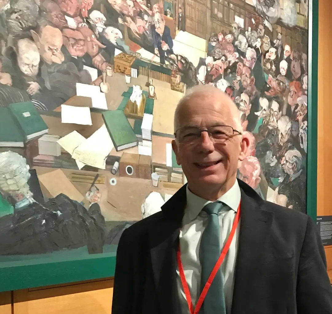Edward Gildea, smiling, in a suit and a red lanyard, in front of a large painting of a crowded debating chamber.