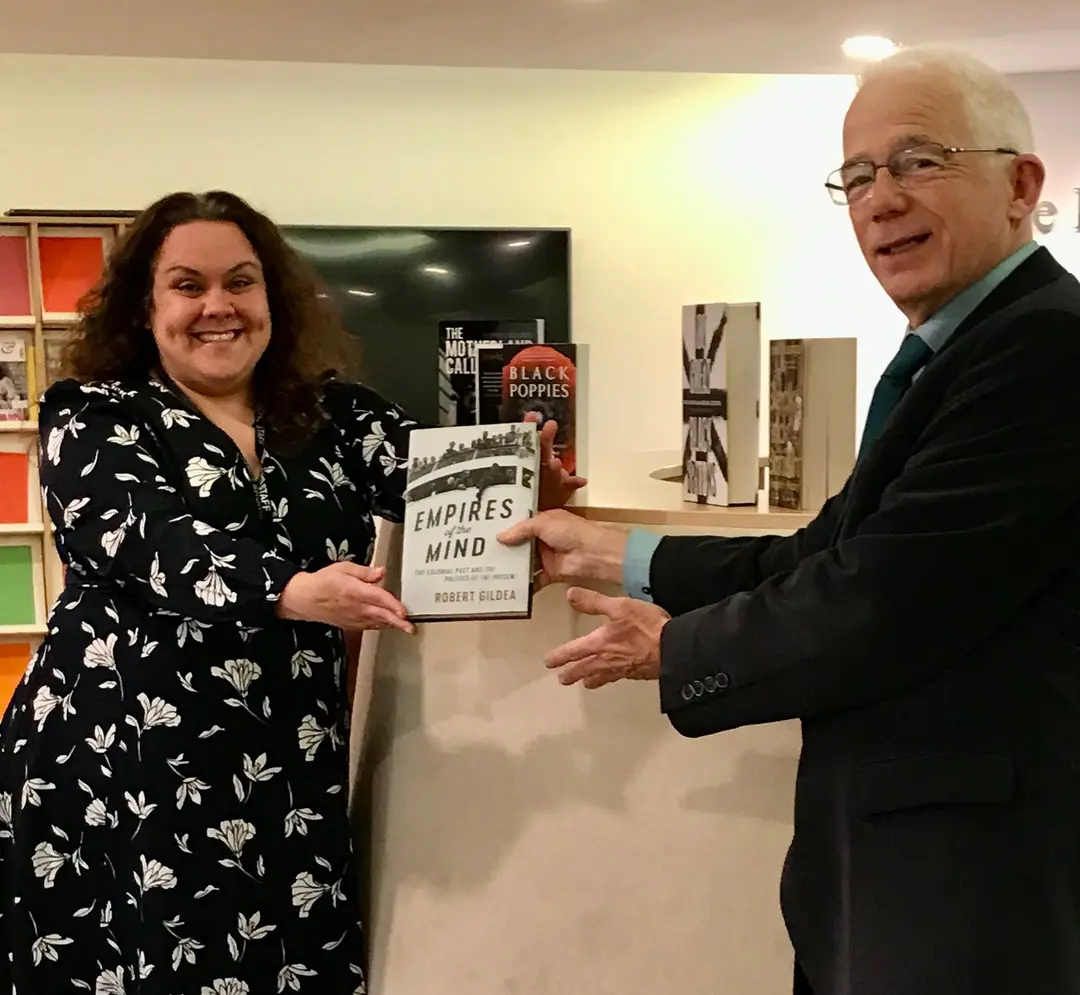 A woman in a floral dress and a man in a dark suit holding a book between them in a library.