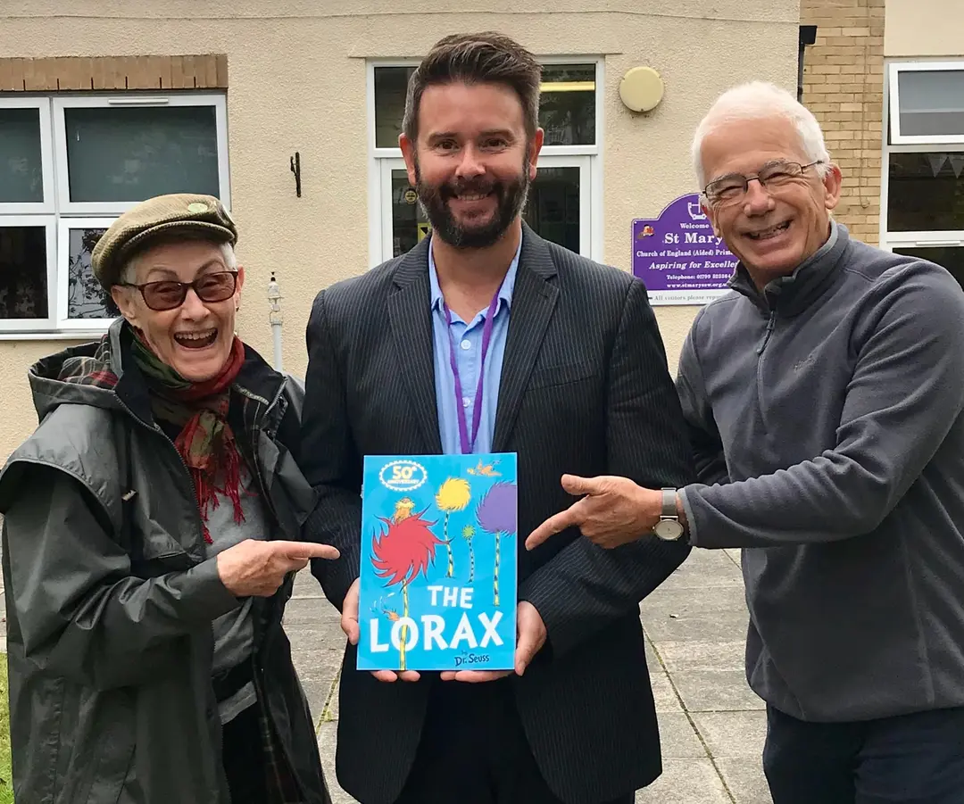 Three smiling people outside a primary school. The man in the middle holds a copy of The Lorax and the two beside him point at it.