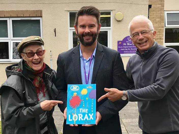 Three smiling people outside a primary school. The man in the middle holds a copy of The Lorax and the two beside him point at it.