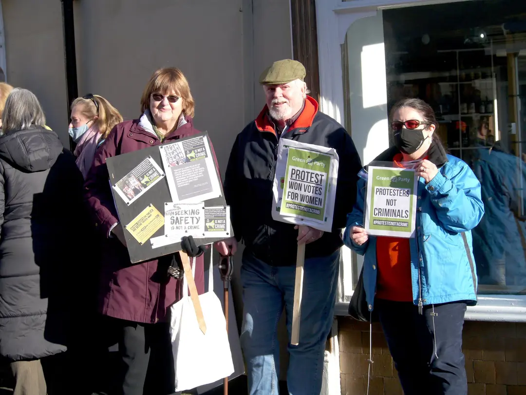 Three people with placards in a sunny street. Two of the placards read “Protest won votes for women” and “Protesters not criminals”.