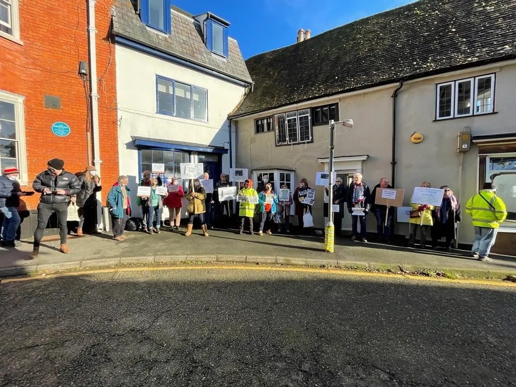 A long line of people holding placards along the pavement in front of a row of buildings, in low winter sun.