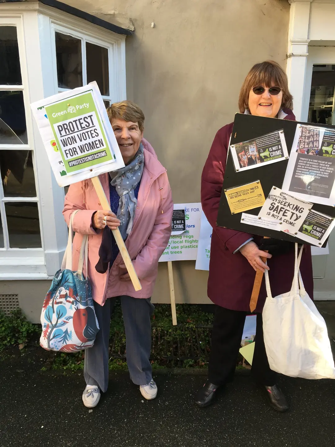 Two women with placards beside a bay window. One placard reads “Protest won votes for women”.