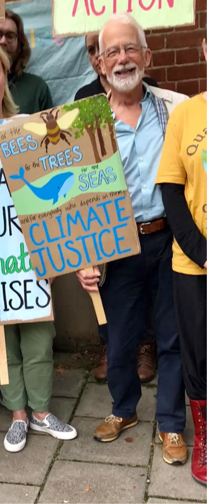 Edward Gildea holding a hand-painted placard that reads “For the bees, for the trees, for the seas, and for everybody who depends on them: climate justice”.