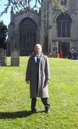 A man in a long grey overcoat standing on the grass in front of a large church.