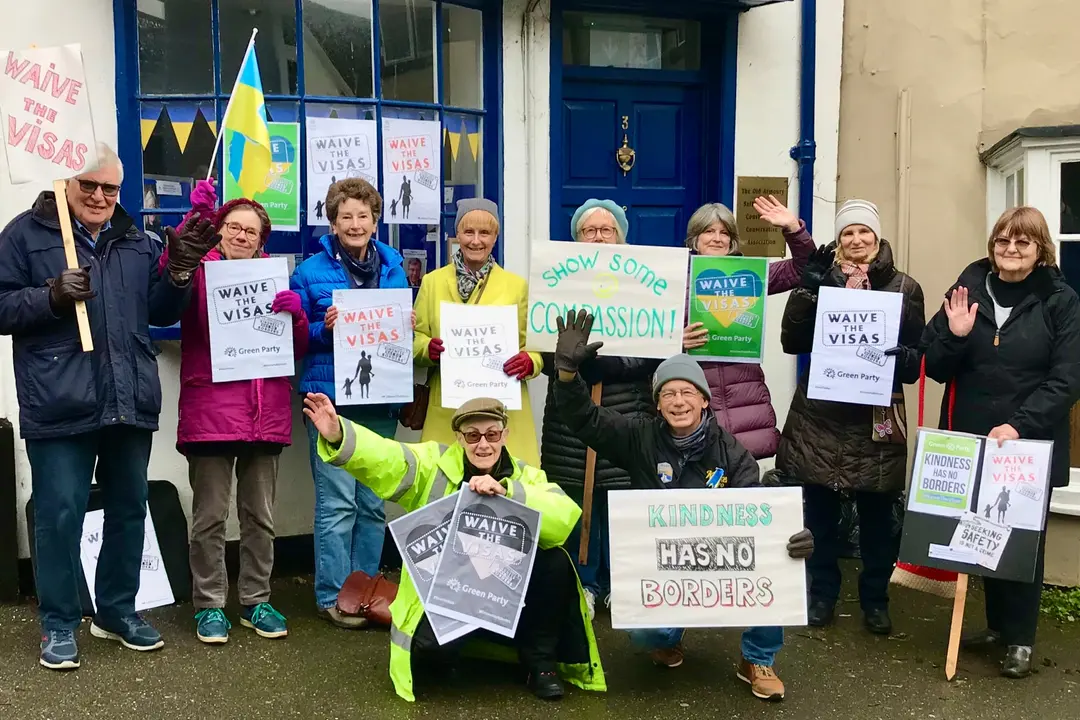 Ten people, smiling and waving, with a Ukrainian flag and placards reading “Waive the visas” and “Kindness has no borders”, outside a building with a blue door.